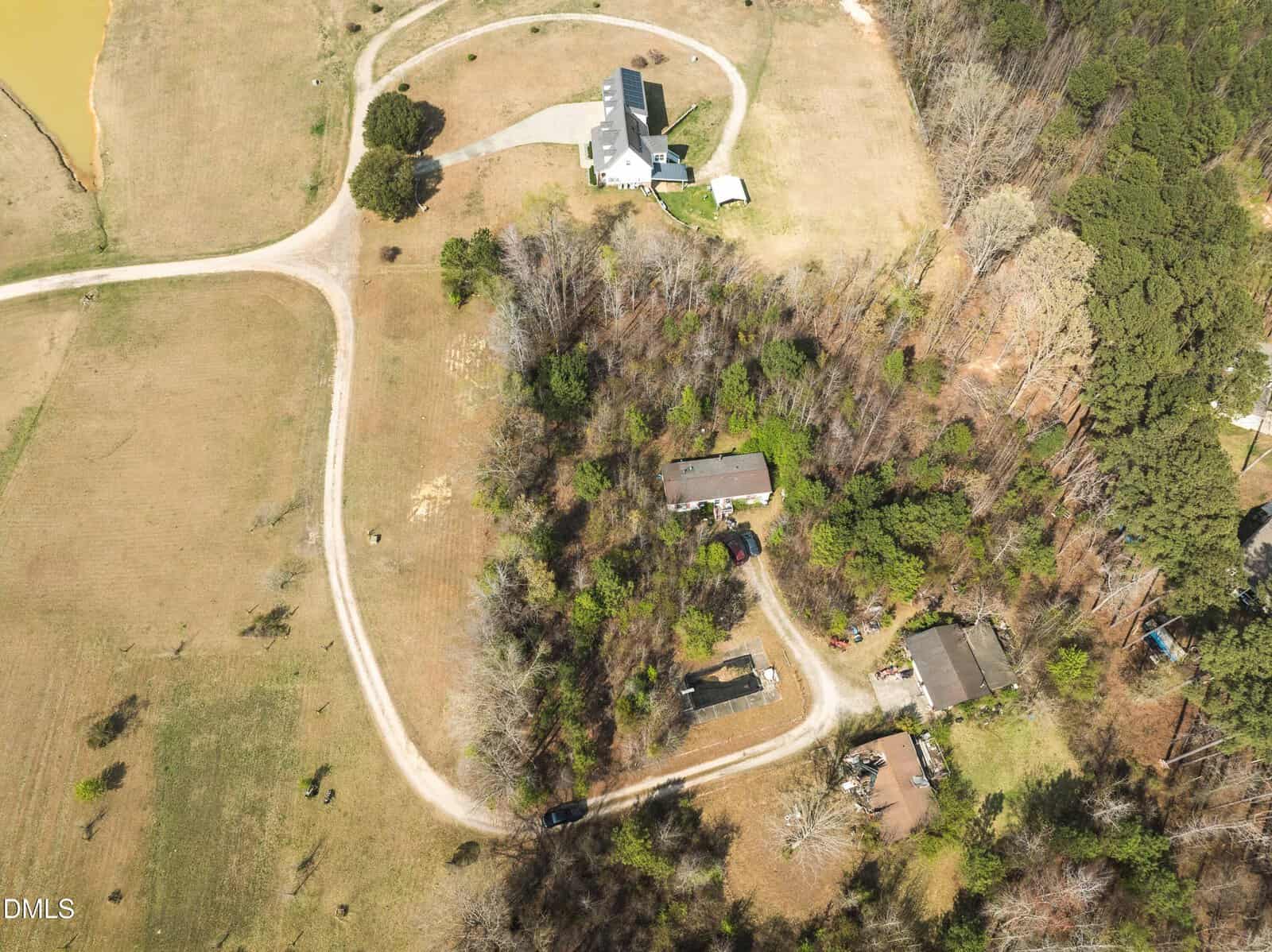 Aerial view of a rural property with a winding driveway, several houses, scattered trees, and open grassy areas next to a pond and forested land.