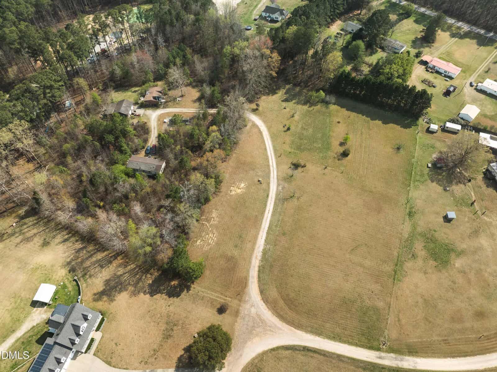 Aerial view of rural homes surrounded by trees and open fields, with a curved dirt driveway leading to several houses and scattered outbuildings under a clear sky.