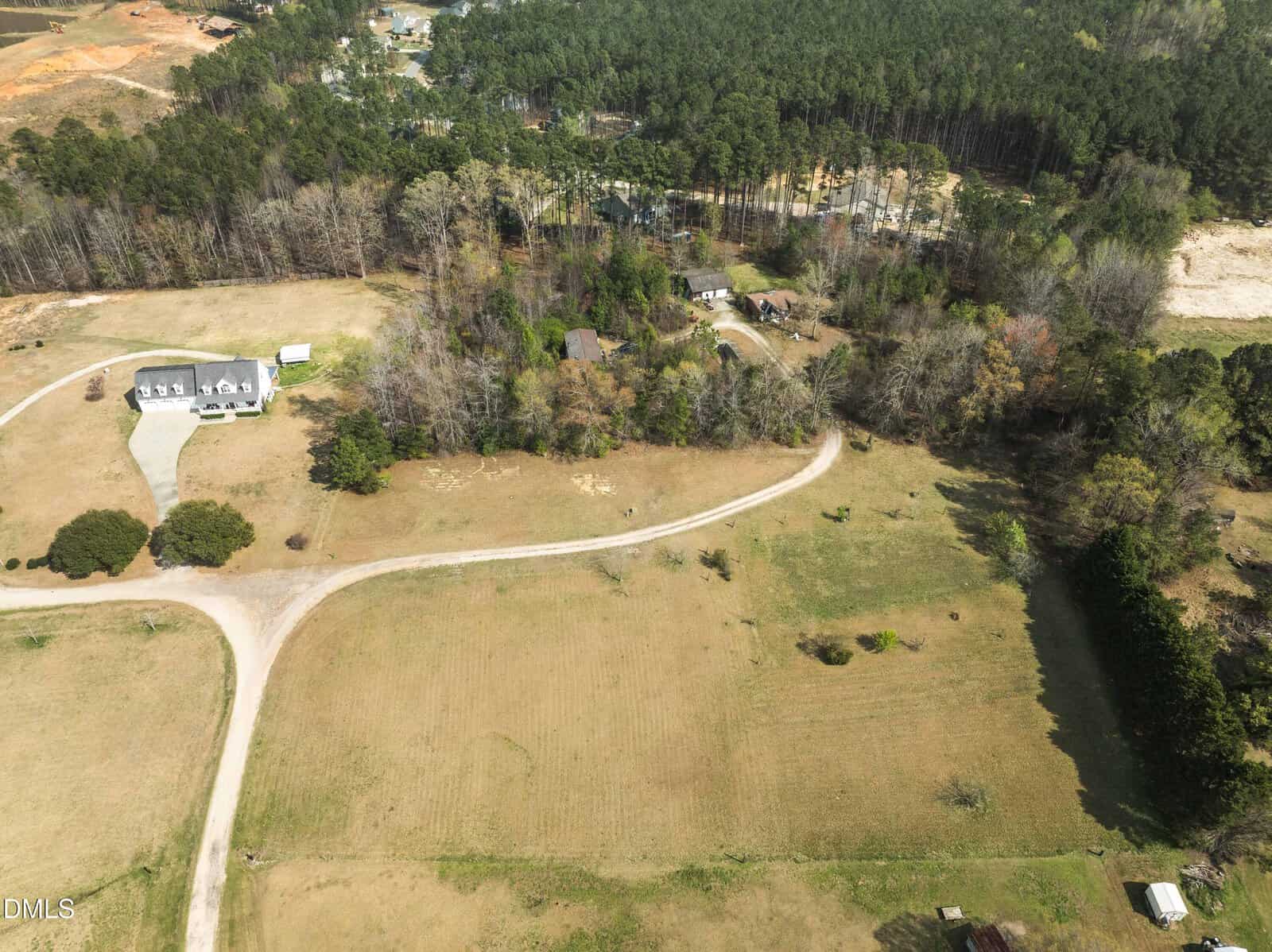 Aerial view of a rural area with a winding dirt road, a large house with a driveway on the left, several trees, smaller buildings in the center and background, and forested land beyond the open fields.