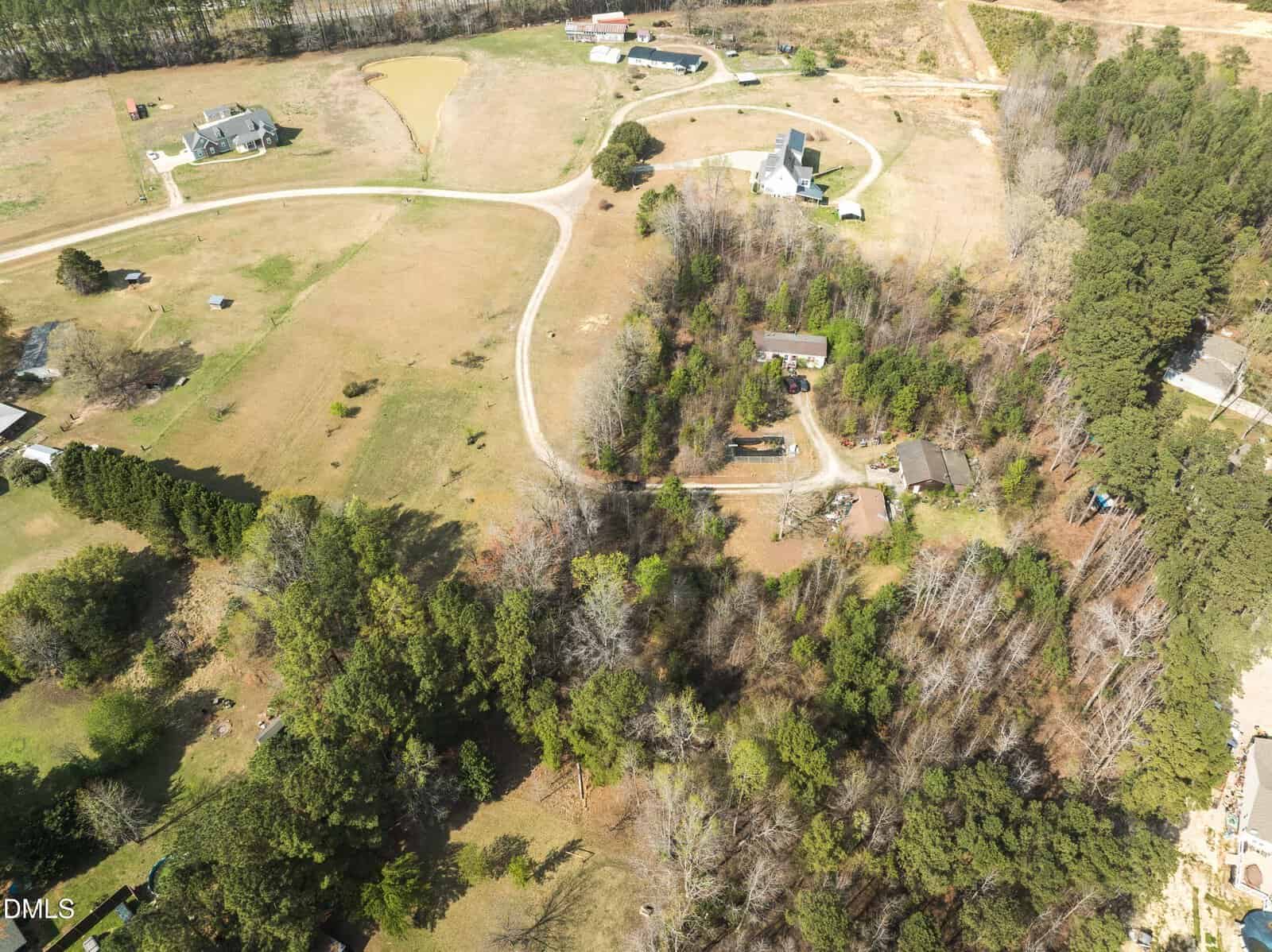 Aerial view of a rural area with several houses, winding driveways, grassy fields, a pond, and clusters of trees. Some buildings are surrounded by wooded areas, and open spaces are visible throughout the landscape.