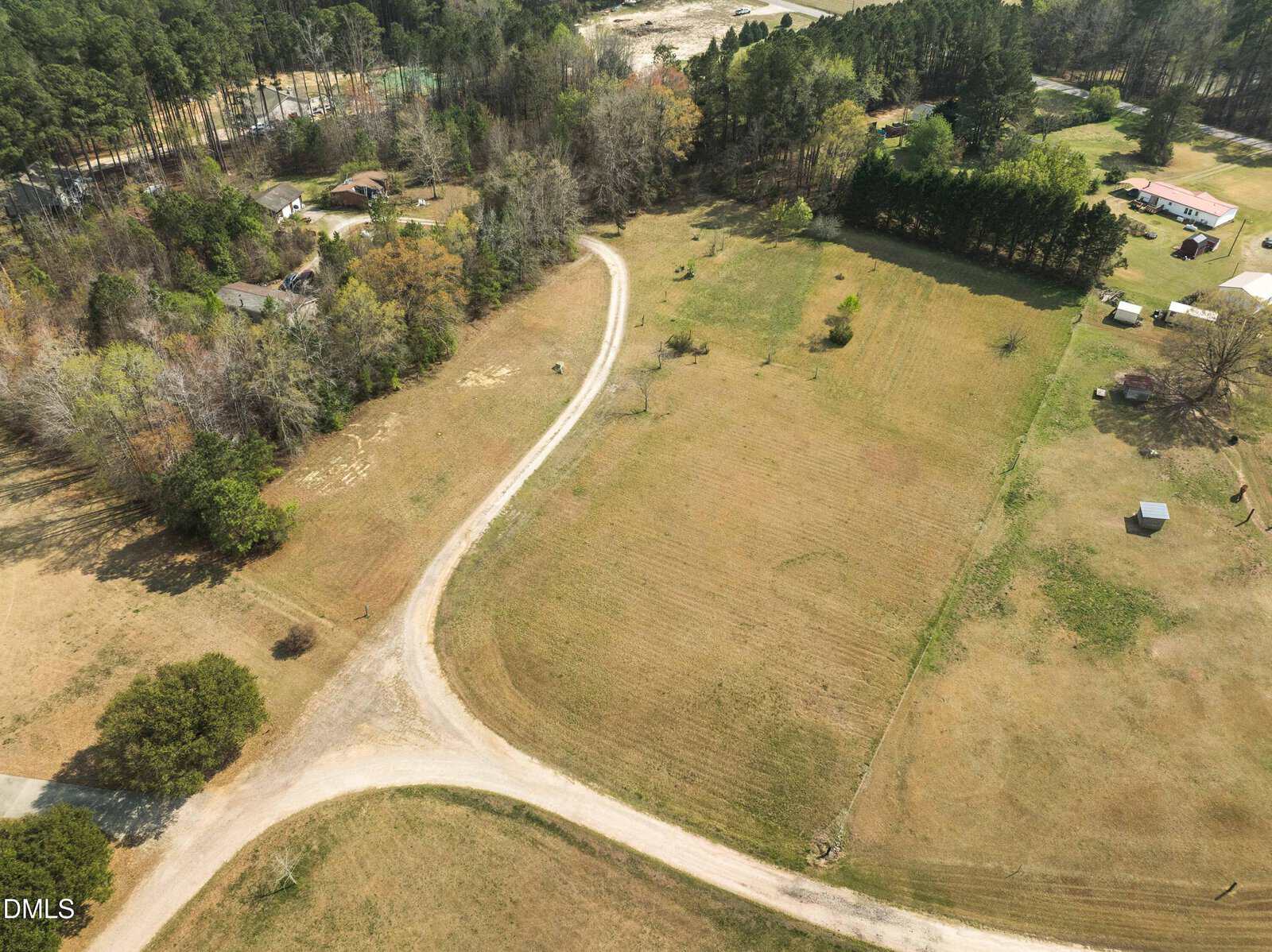 Aerial view of a rural area with houses surrounded by trees, open grassy fields, and a winding dirt road running through the landscape.