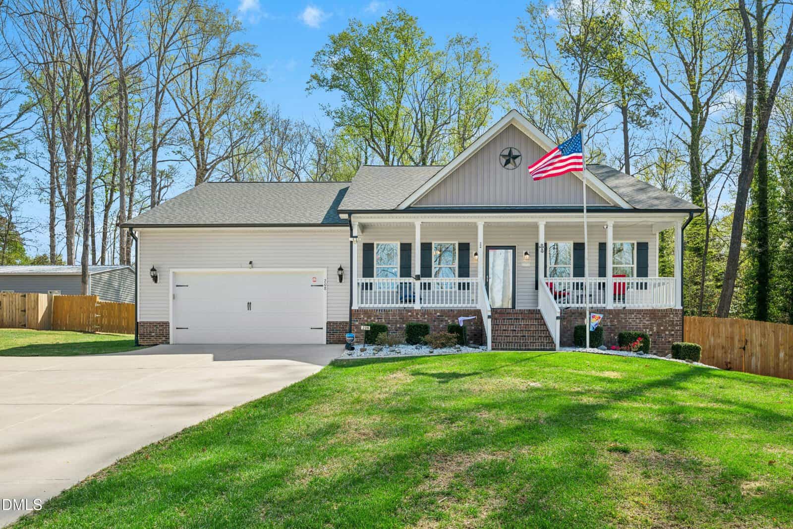 A single-story house with a front porch, brick and siding exterior, attached two-car garage, American flag on a pole, and a well-kept lawn, surrounded by a wooden fence and trees in the background.