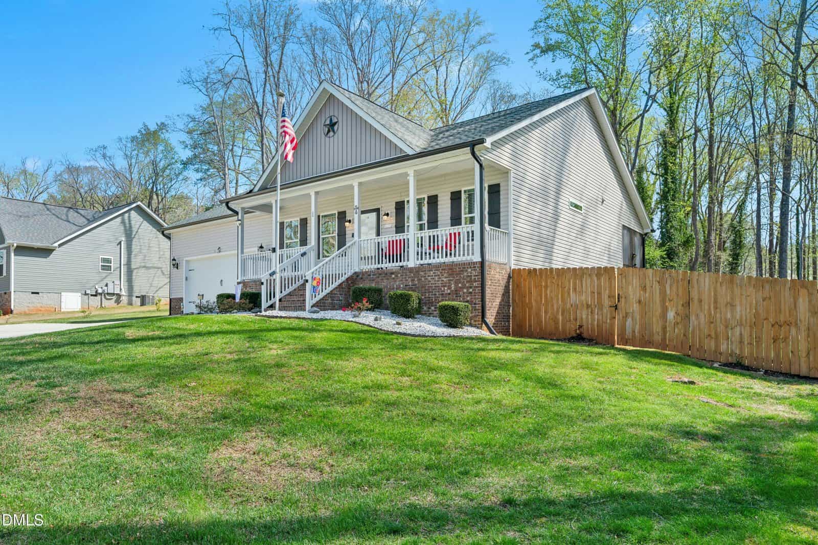 Single-story house with light gray siding, a covered front porch, an American flag, white railing, and brick accents. The yard is grassy with mulch beds, and a wooden fence runs along the right side of the property.