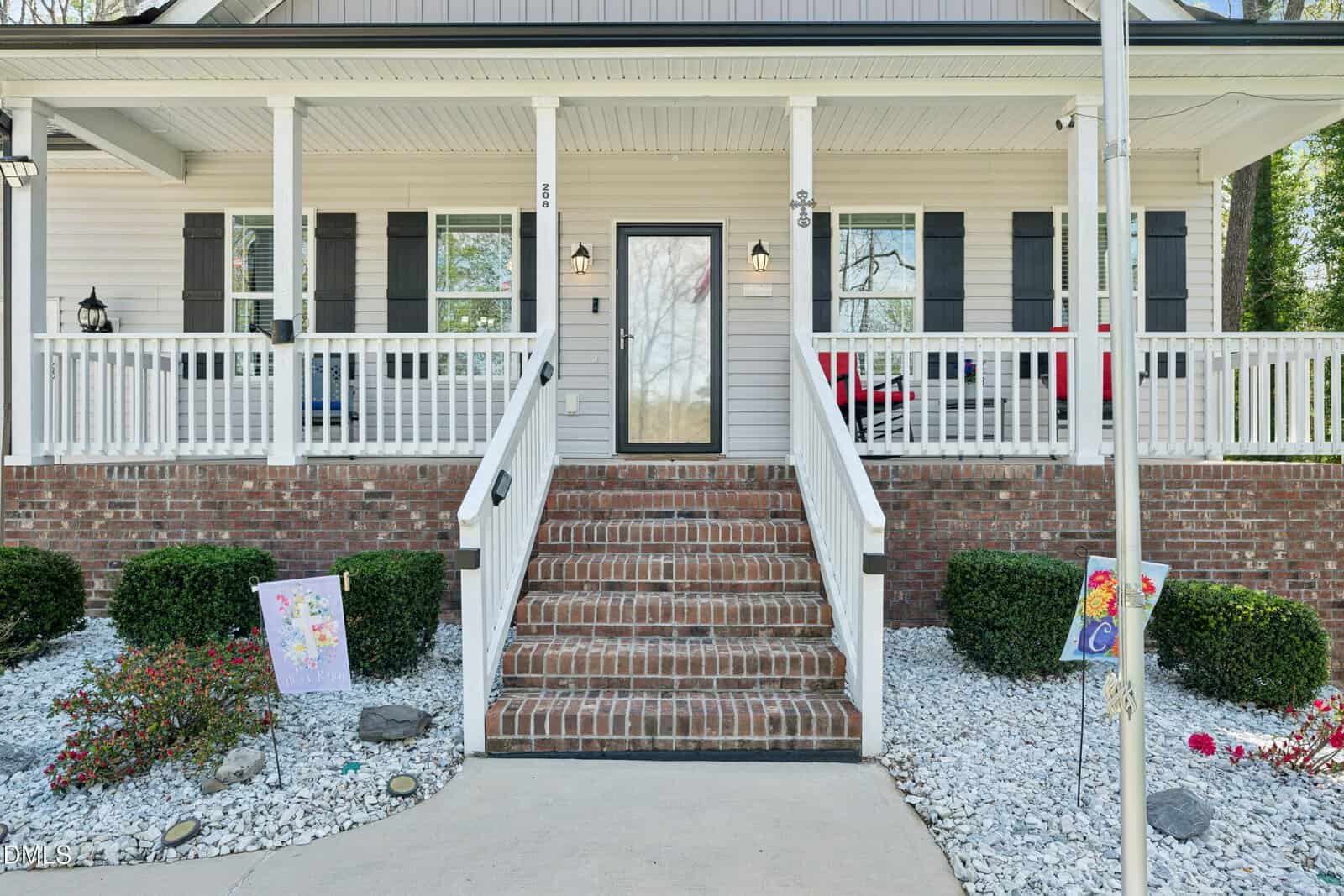 Front view of a house with brick steps leading to a glass-paneled door, white railing, white siding, black shutters, small bushes, white gravel, and two small decorative flags in the landscaped yard.