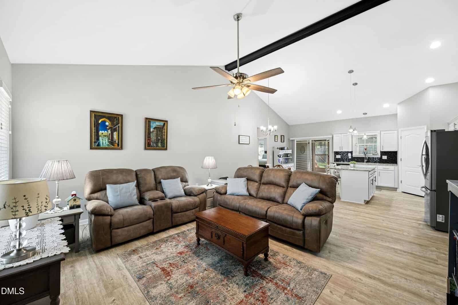 A cozy living room with two brown sofas, a wooden coffee table, floor lamp, artwork on the wall, and view into a kitchen with white cabinets and a ceiling fan overhead.