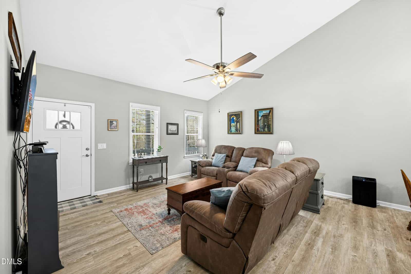 A living room with light gray walls, wood flooring, a brown sectional sofa, a wooden coffee table, a TV mounted on the wall, and several framed pictures. A ceiling fan with lights hangs from the vaulted ceiling.