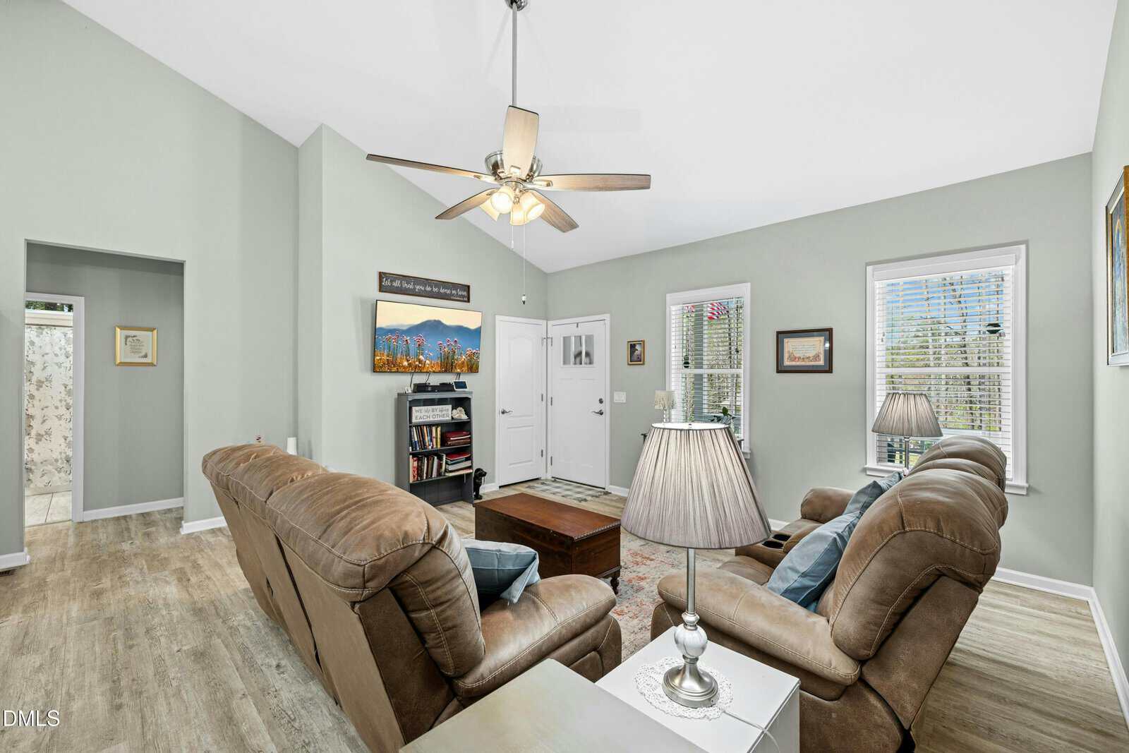 Bright living room with two brown recliners, a couch, a wooden coffee table, two lamps, wall art, bookshelves, light gray walls, wood floors, and windows letting in natural light. Door and hallway visible in the background.
