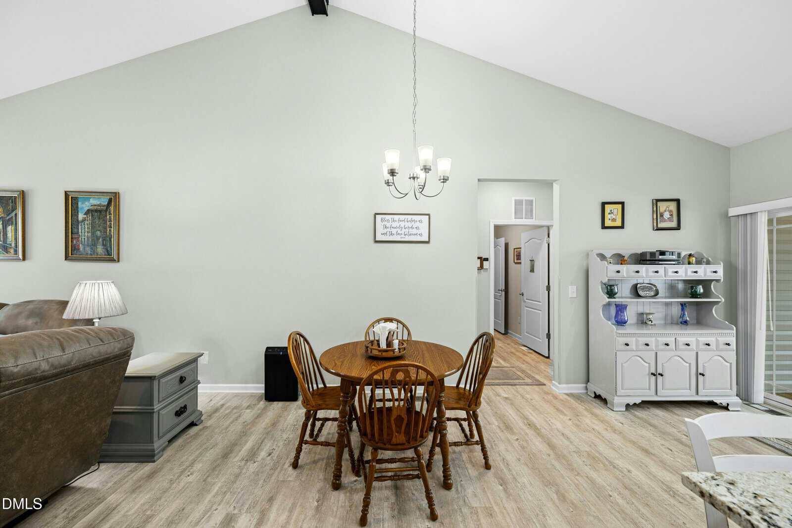 Dining area with a round wooden table and four chairs, chandelier above, white walls, wood flooring, and a sideboard with decor. A living area and kitchen are partially visible.