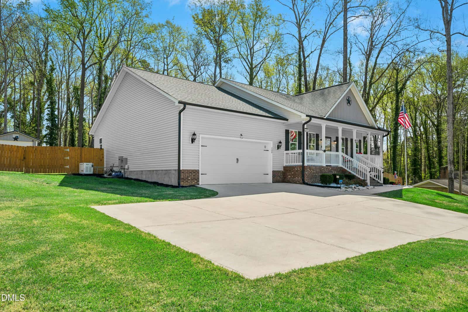 A single-story, white house with a gray roof, attached garage, front porch with white railings, and an American flag. The house sits on a large lawn bordered by trees, with a wide concrete driveway.