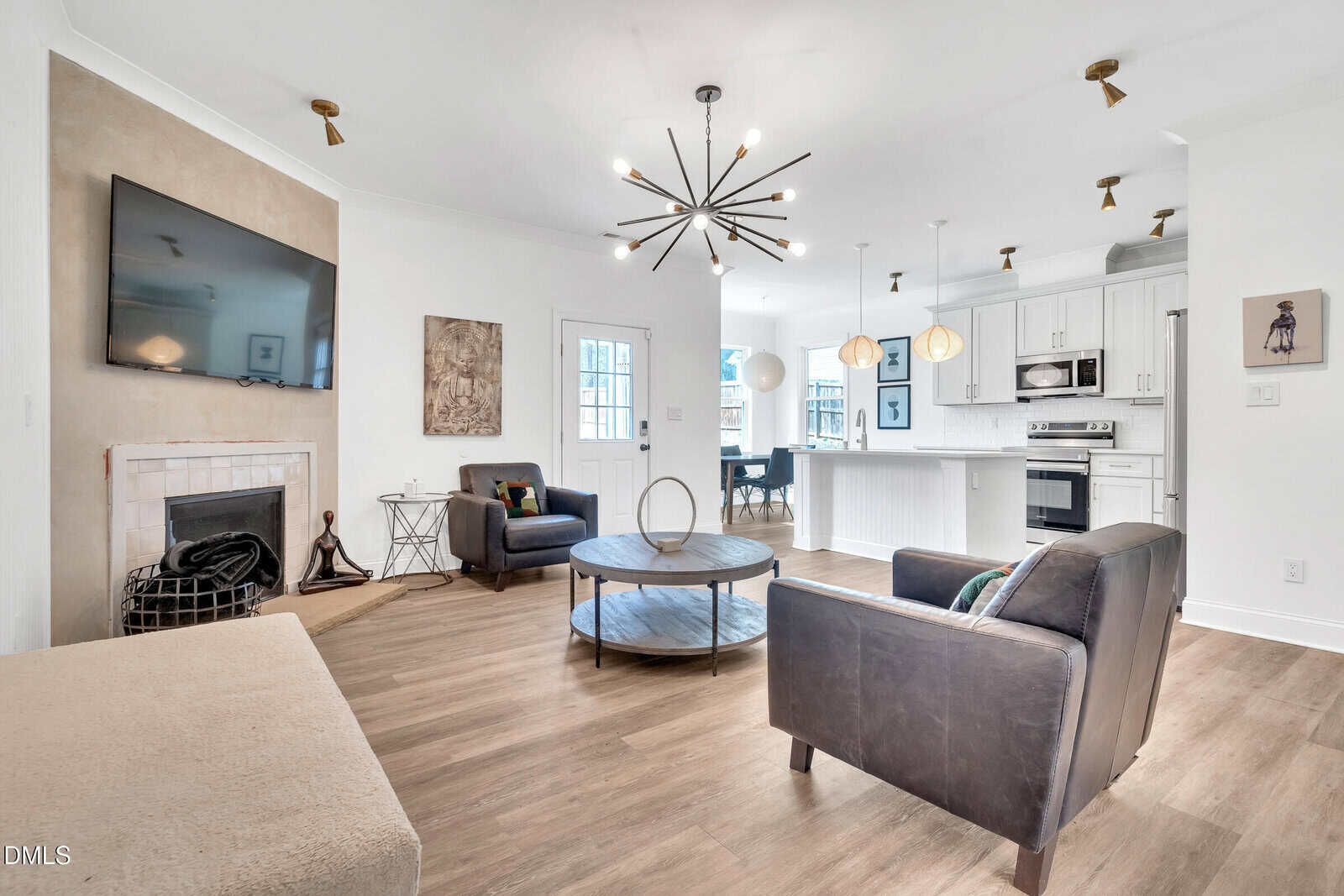 Modern living room with wood flooring, two armchairs, wall-mounted TV, round coffee table, fireplace, and view into a bright white kitchen with island seating and pendant lights. Art and decor add warmth to the space.