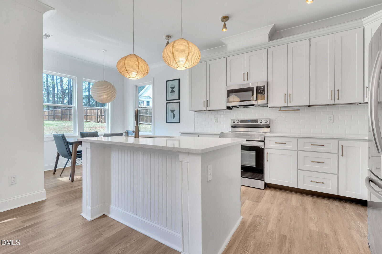 A modern kitchen with white cabinets, stainless steel appliances, an island with a white countertop, and two round pendant lights. A dining area with large windows and a table is visible in the background.