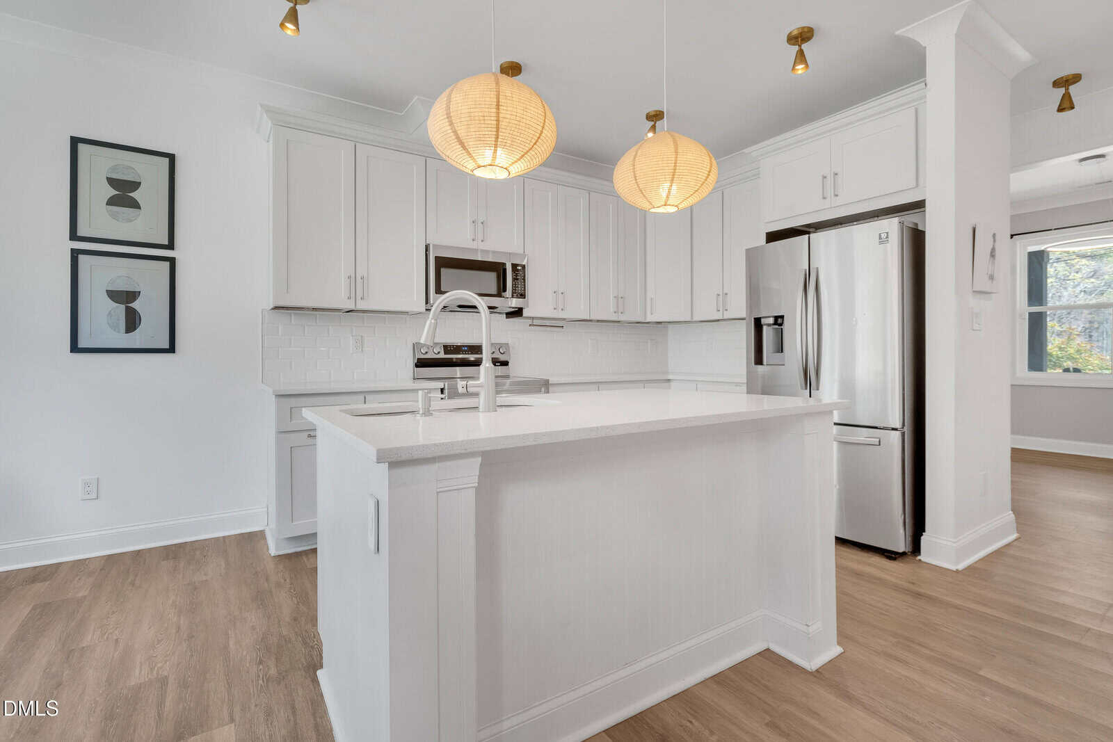 A modern kitchen with white cabinets, stainless steel appliances, a white island with a sink, two round pendant lights, light wood flooring, and two abstract framed prints on the wall.