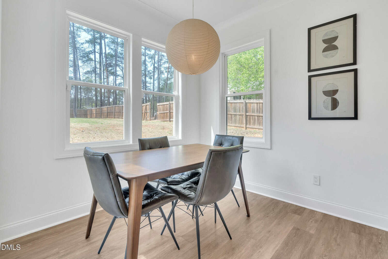 A wooden dining table with four gray cushioned chairs sits by two large windows. A round pendant light hangs above, and two abstract art pieces are on the white wall. A fenced backyard is visible outside.