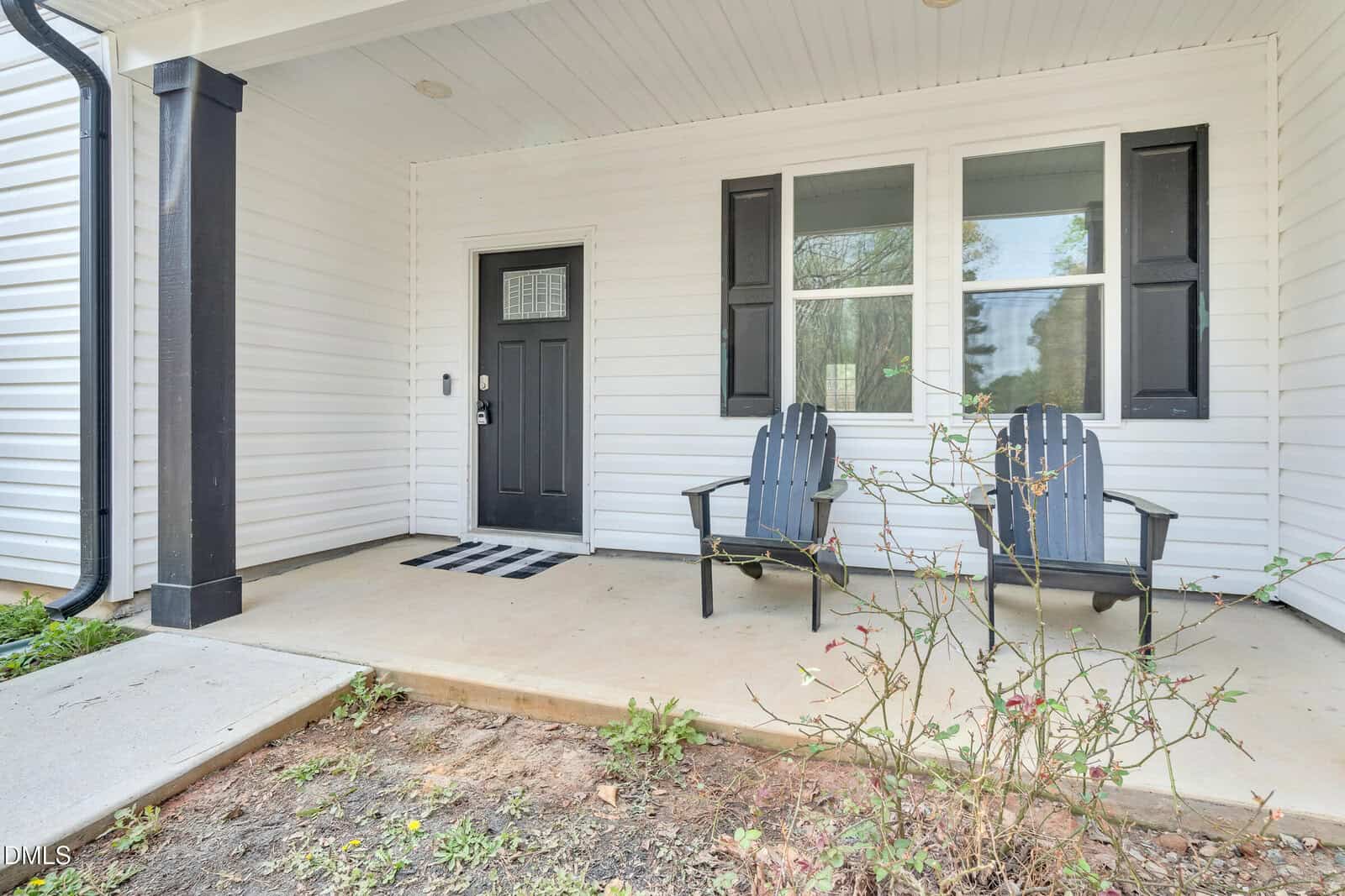 A covered front porch with two black Adirondack chairs, a black front door, two large windows with black shutters, and a small garden area with a few plants in front.