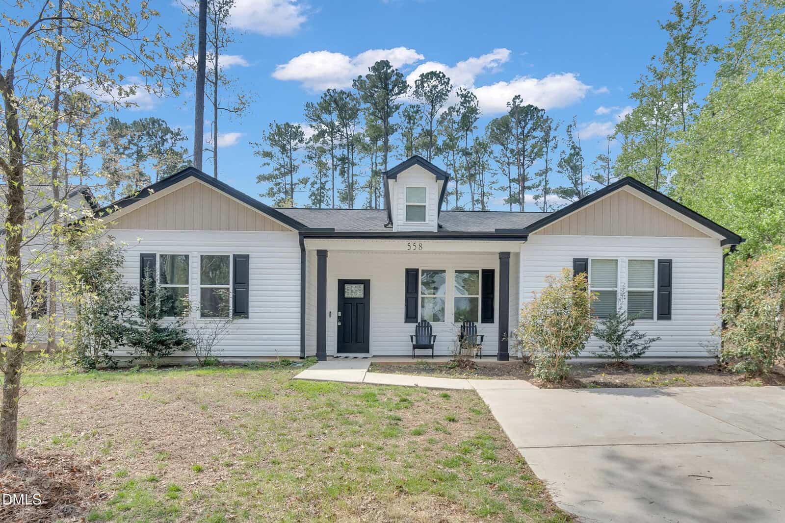 Single-story white house with black trim and a central dormer, featuring a black front door, two chairs on the small porch, and a driveway on the right. Tall trees and shrubs surround the home under a partly cloudy sky.