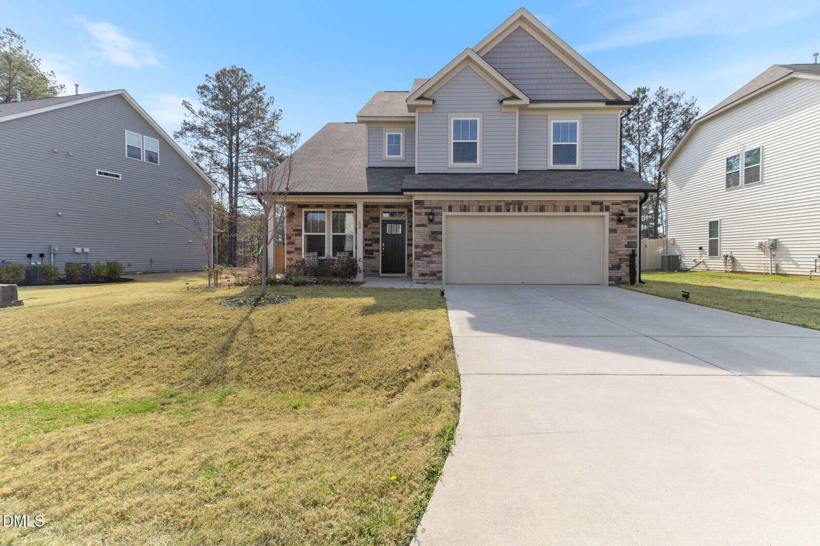 Two-story suburban house with a gray exterior, attached two-car garage, and stone accents. Large driveway, well-kept lawn, and neighboring houses on both sides under a clear blue sky.