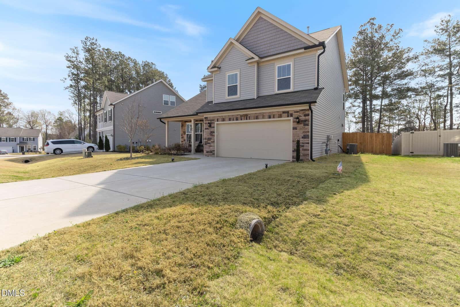 Two-story suburban house with a gray exterior, stone accents, double garage, and large driveway. The lawn is neatly trimmed, and trees surround the property under a clear blue sky.