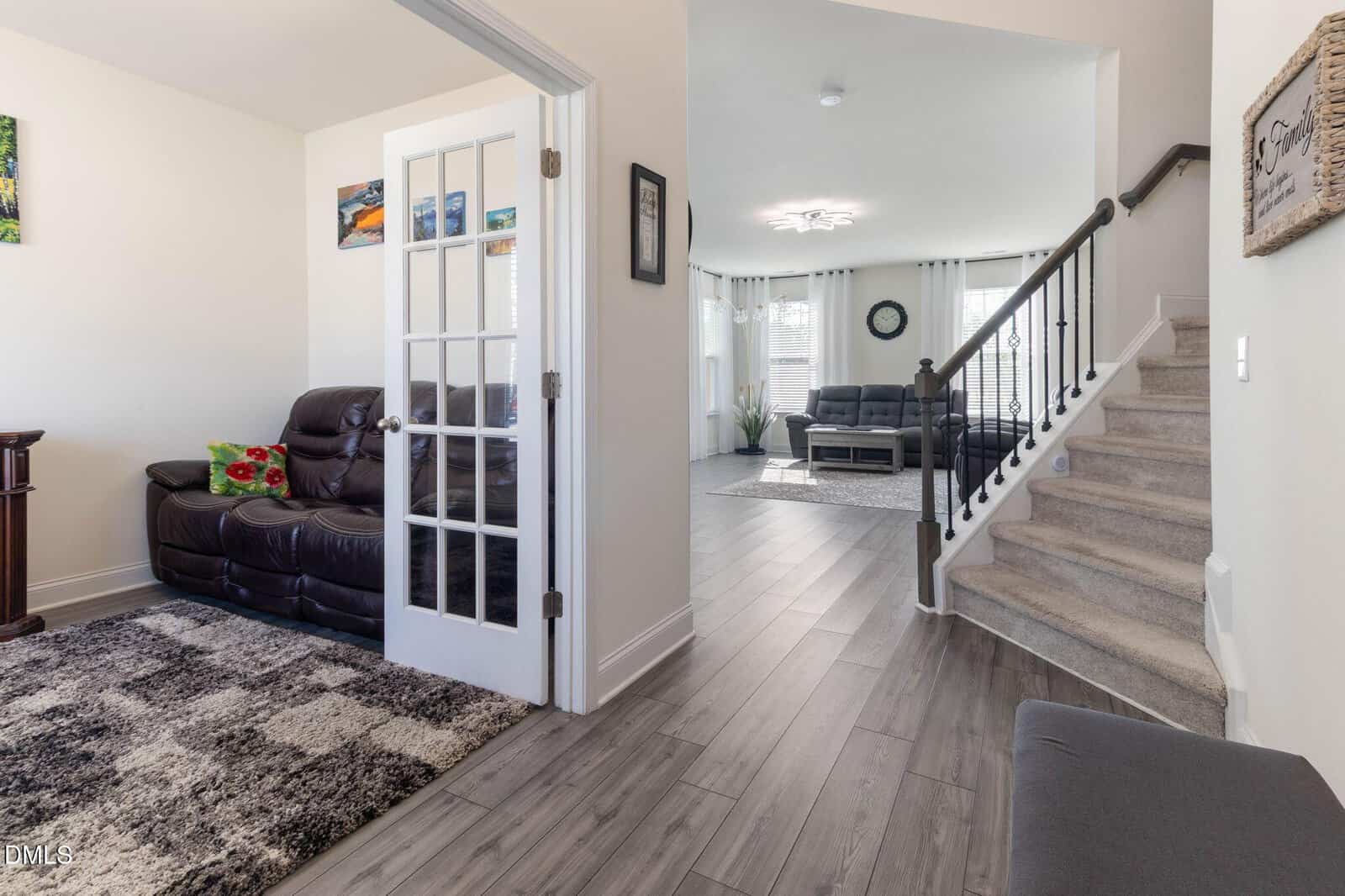 A modern home interior with a staircase, a living area with brown leather sofas, large windows, a glass-paned door, and wood-look tile flooring. Bright and spacious, decorated in neutral tones with accents and wall art.