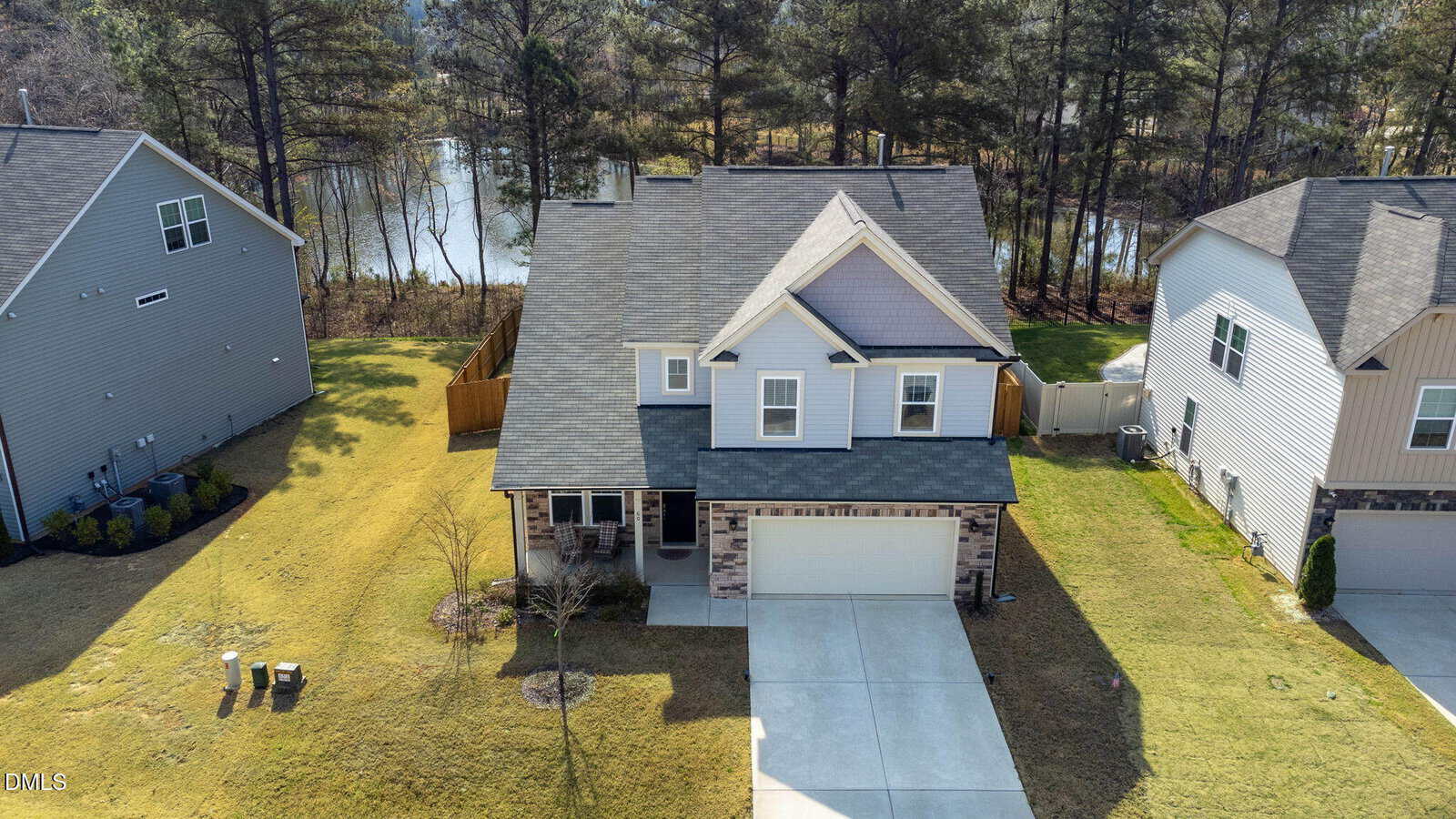 A two-story suburban house with a gray roof, light-colored siding, and stone accents. The home has a front porch, driveway, and a fenced backyard. Trees and a lake are visible in the background.
