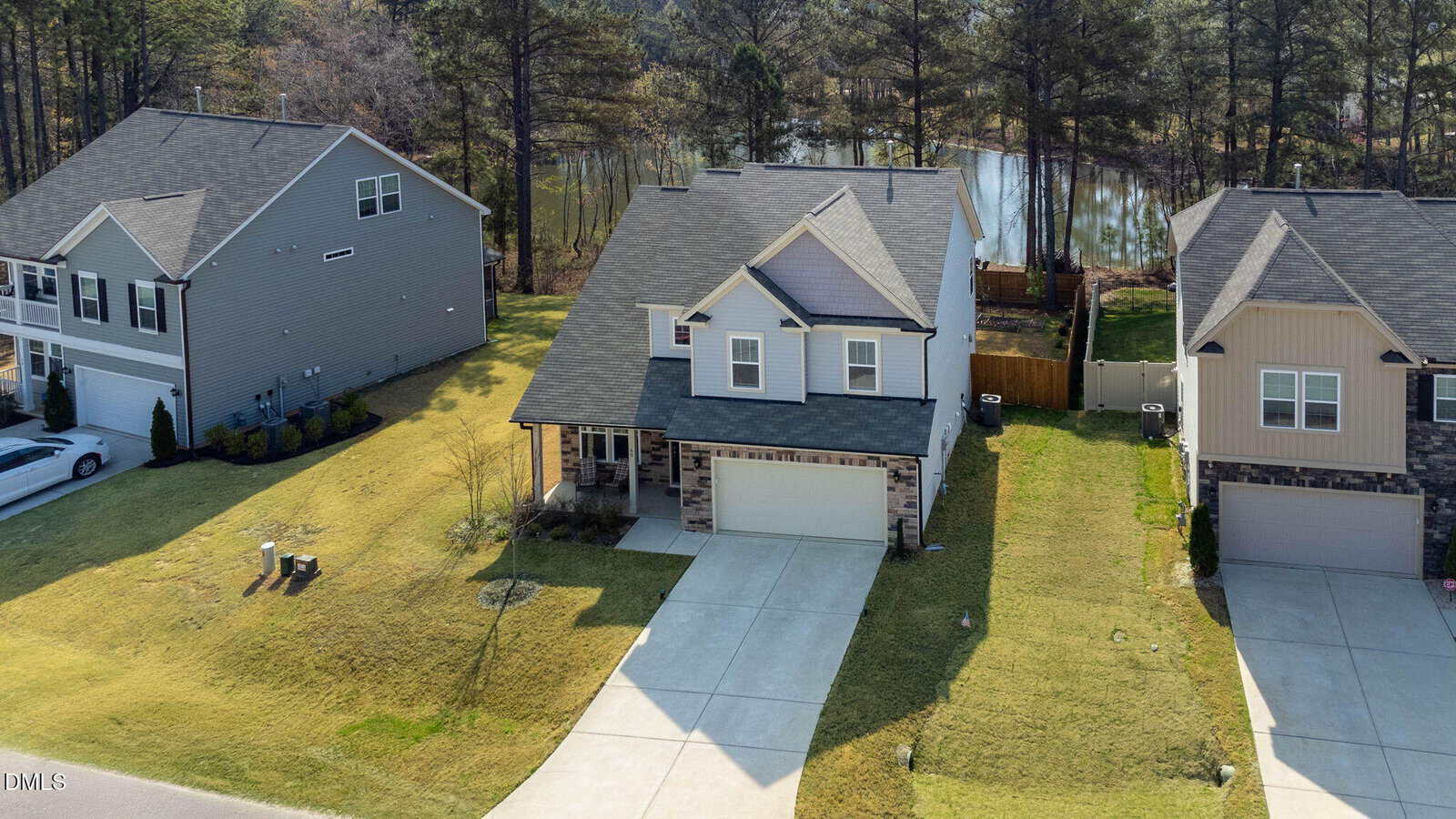 A two-story suburban house with a covered front porch, attached two-car garage, and stone accents, situated between two similar houses, with a backyard leading to trees and a small lake.