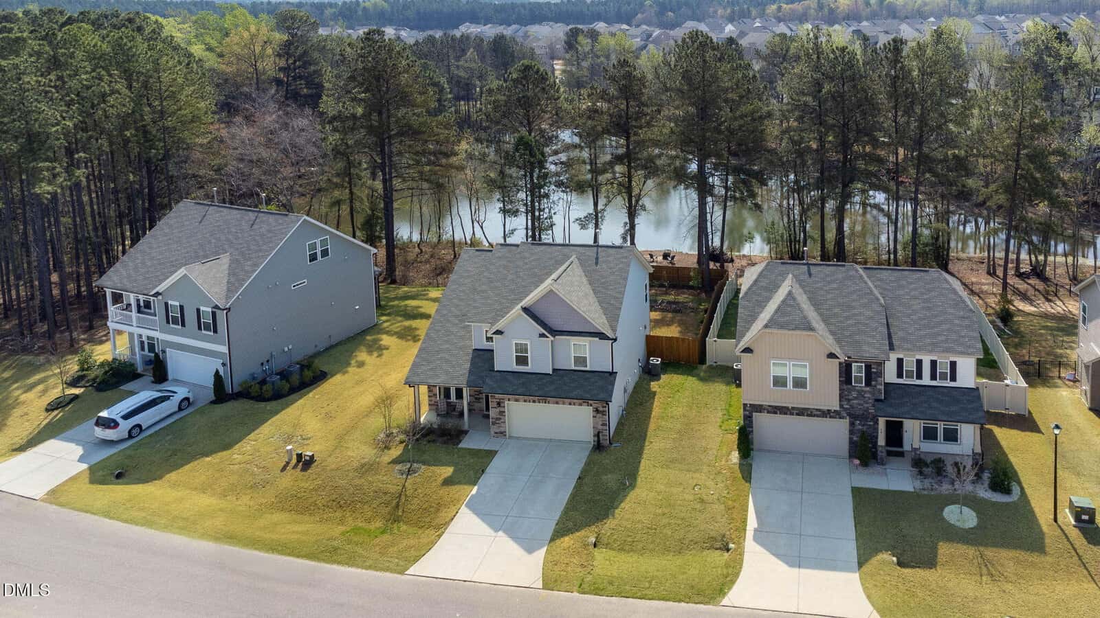 Aerial view of three two-story suburban houses with driveways and garages, situated on a quiet street. Behind the houses is a wooded area and a pond. The lawns are neatly maintained and a white car is parked in one driveway.