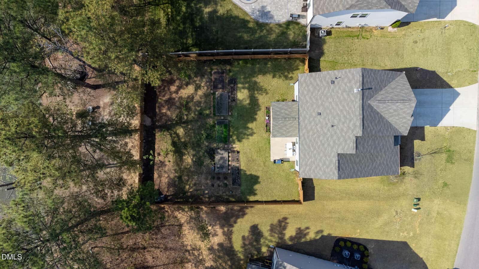 Aerial view of a suburban house with a fenced backyard containing two rectangular garden beds, a small patio, and surrounding trees casting shadows on the lawn. The driveway and part of the street are visible.