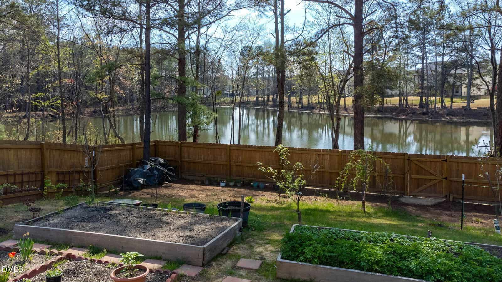 A fenced backyard with raised garden beds, a few young trees, and a lawn overlooks a wooded pond. Gardening supplies and pots are near the fence, and tall trees surround the pond in the background.