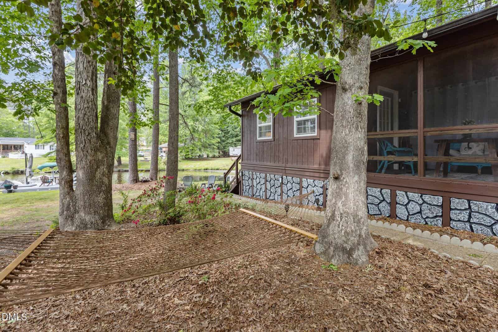 A wooden cabin with a screened porch sits among trees, overlooking a lake. A hammock is stretched between two trees in the foreground, with a garden area and dock visible near the water in the background.