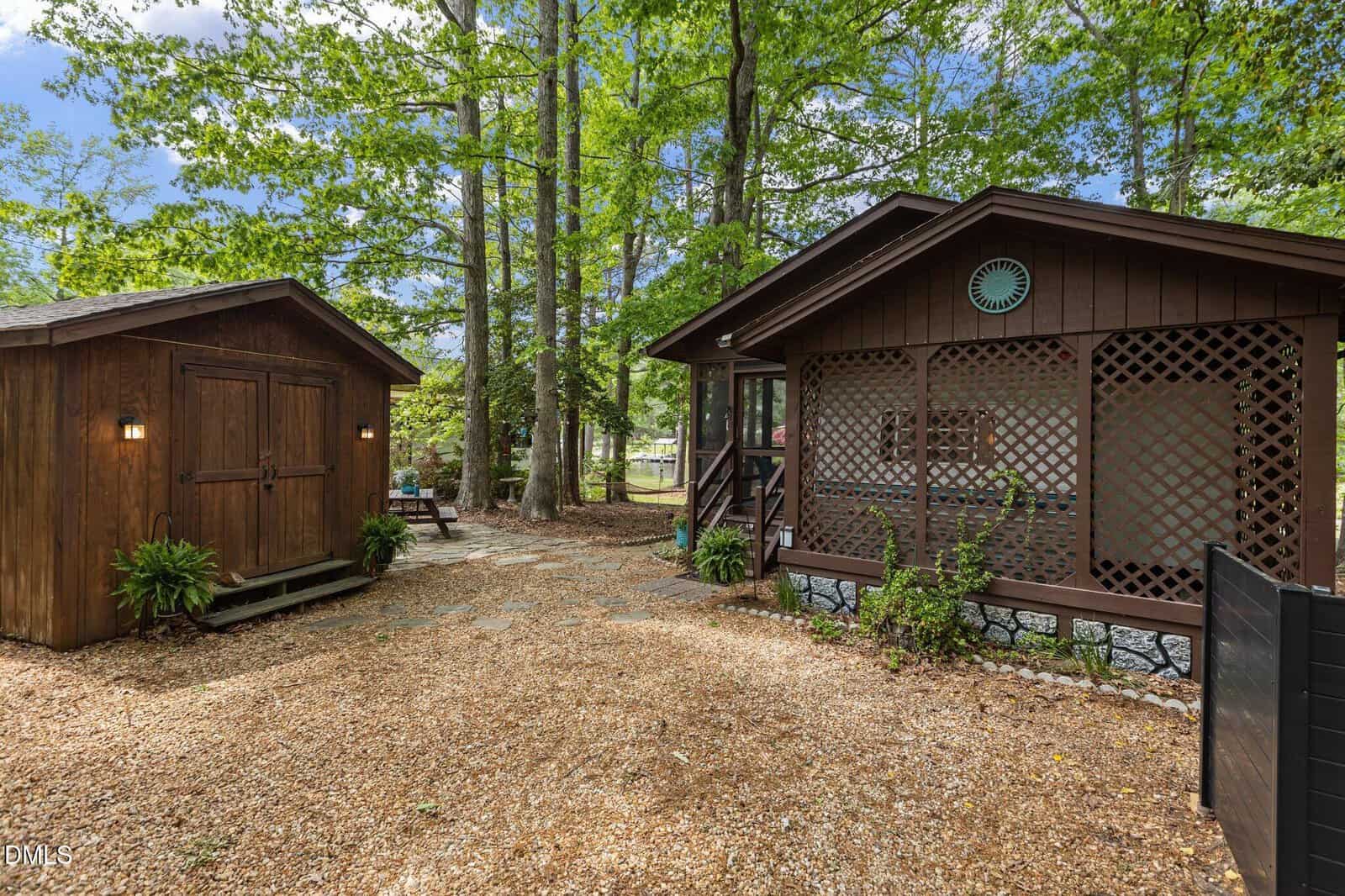 A cozy brown cabin with a screened porch sits among tall trees, next to a matching wooden shed. A gravel path and greenery surround the buildings, creating a peaceful, rustic outdoor setting.