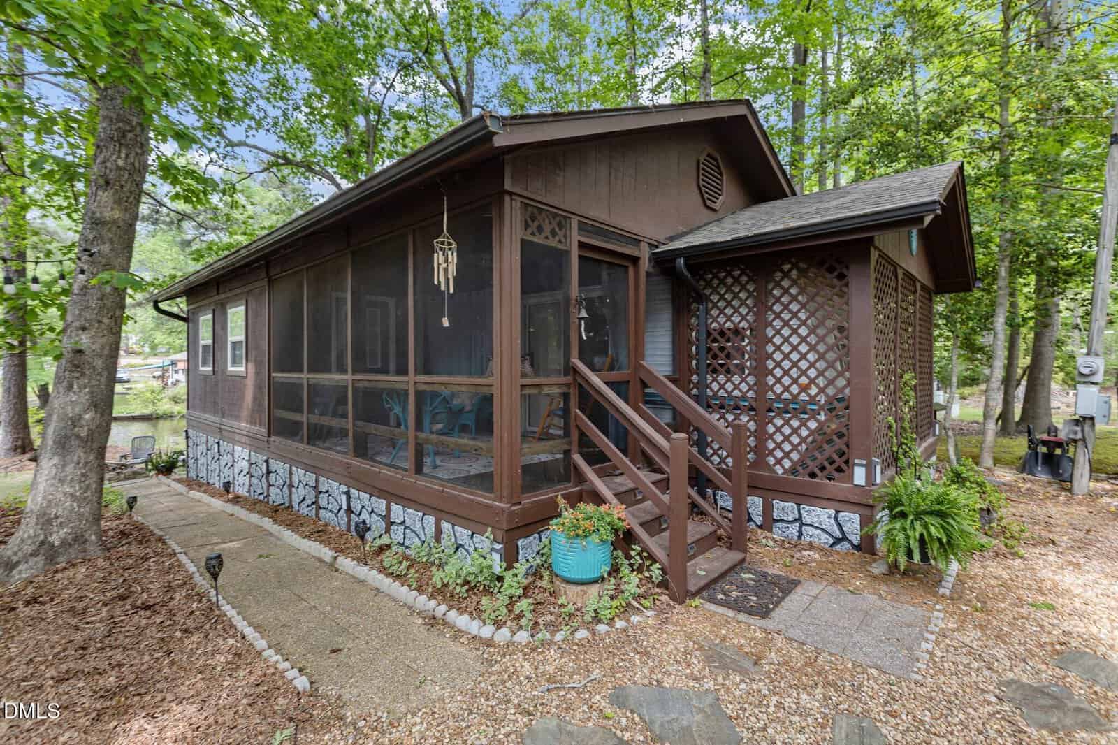 A small brown cabin at 106 Desoto Drive, Louisburg, features a screened-in porch nestled among tall trees. Potted plants, a wind chime, and a stone walkway welcome you to the mulch-covered yard bordered with white stones.
