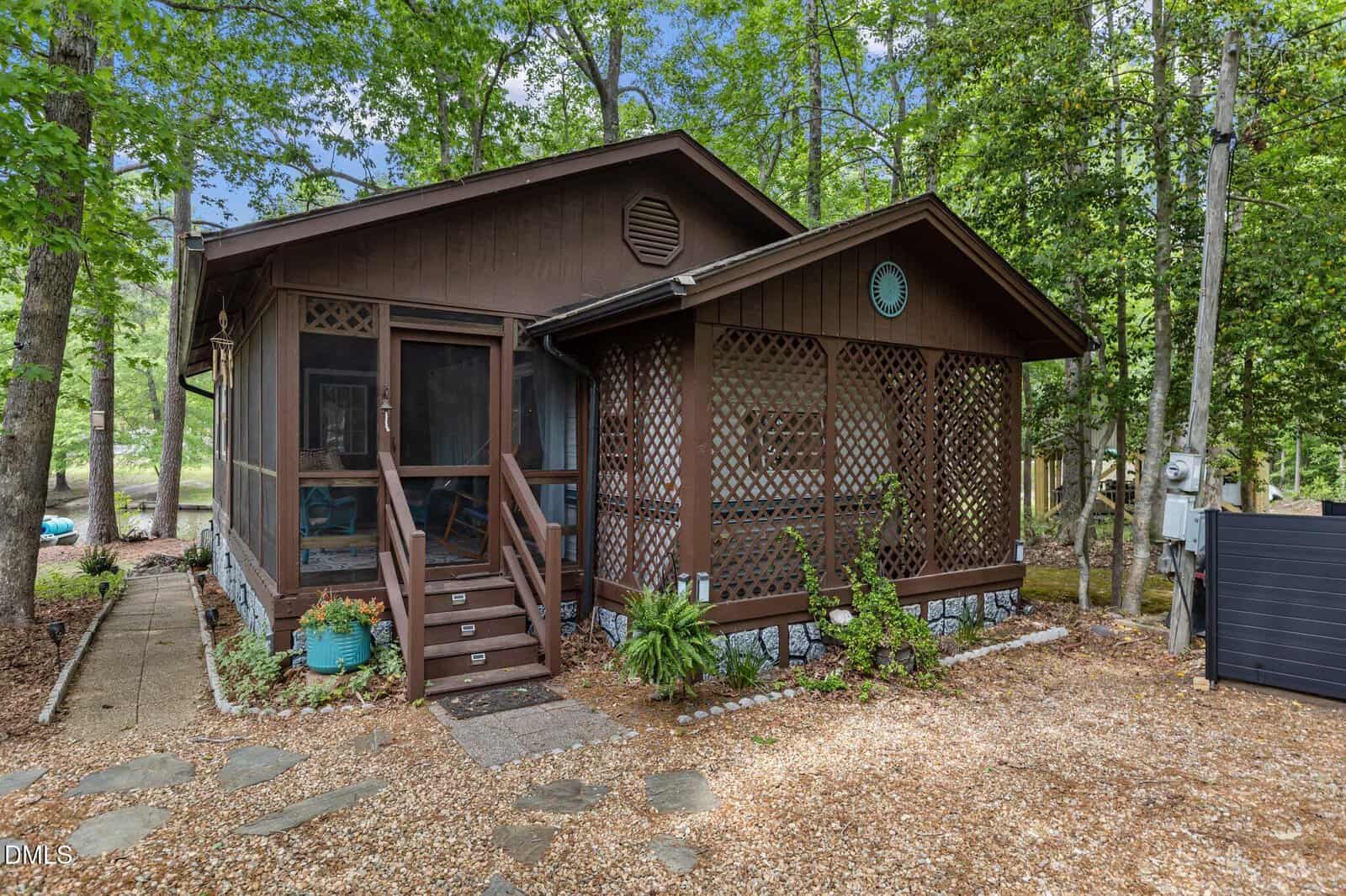 A small brown cabin with lattice woodwork and a screened porch, surrounded by tall green trees and gravel walkways, set in a wooded area. Steps and potted plants decorate the entrance.