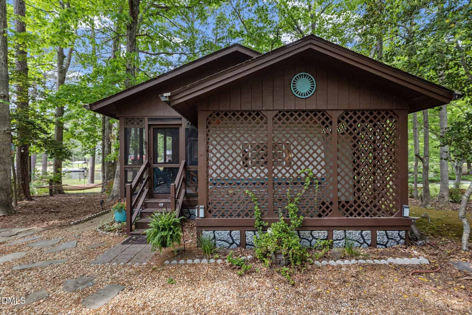 A small brown wooden cabin with a front porch, lattice details, and a circular decorative piece above the entrance, surrounded by trees and a gravel pathway in a wooded area.