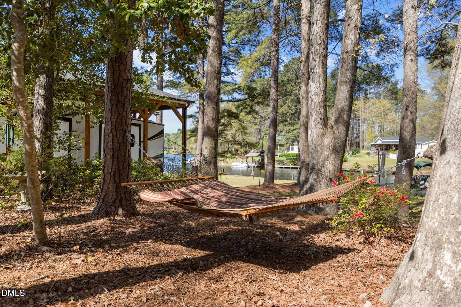 A striped hammock is tied between trees in a wooded yard with nearby houses, flowering bushes, and boats visible in the background on a sunny day.