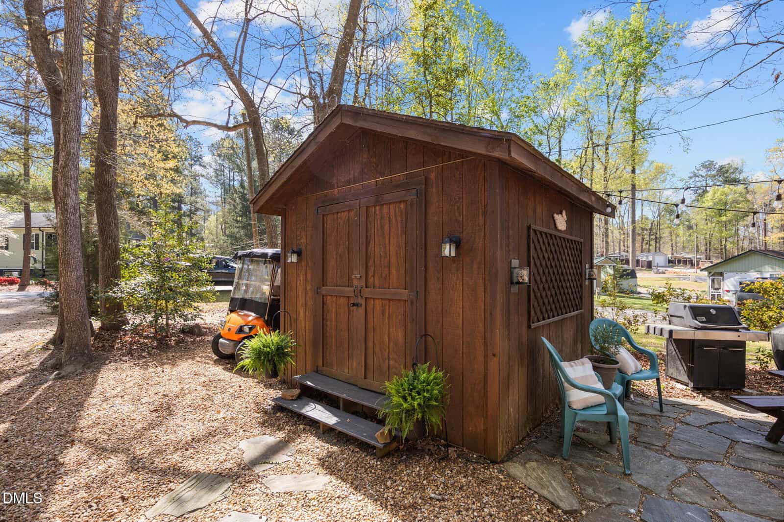A small wooden shed with double doors sits among trees. Two chairs and potted ferns are nearby, with a golf cart and parked vehicles visible in the background on a sunny day.