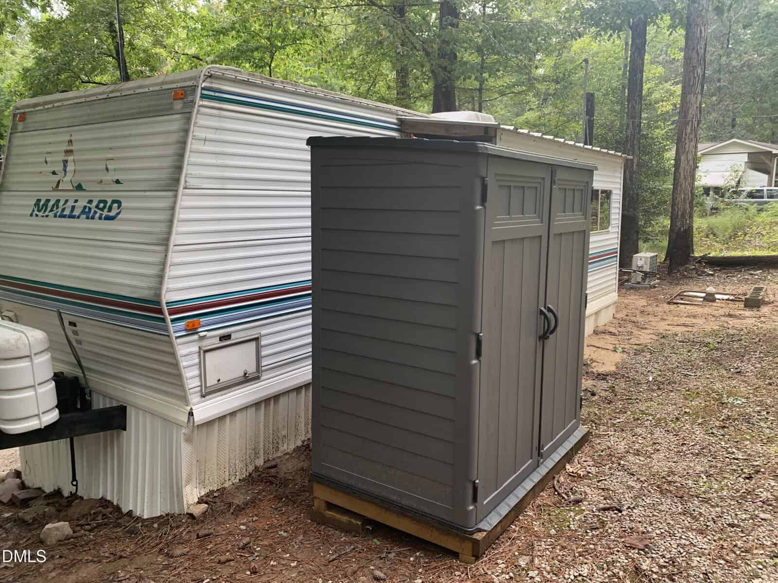 A gray outdoor storage shed with double doors stands on a wooden platform next to a white Mallard travel trailer in a wooded area with trees and a house visible in the background.