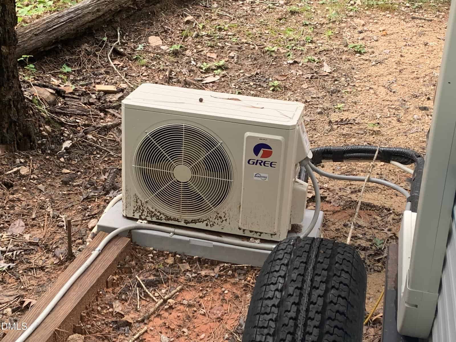An outdoor Gree air conditioning unit sits on a concrete pad surrounded by dirt, leaves, and some grass, with several cables and pipes connected to it. A tire and wooden plank are visible in the foreground.