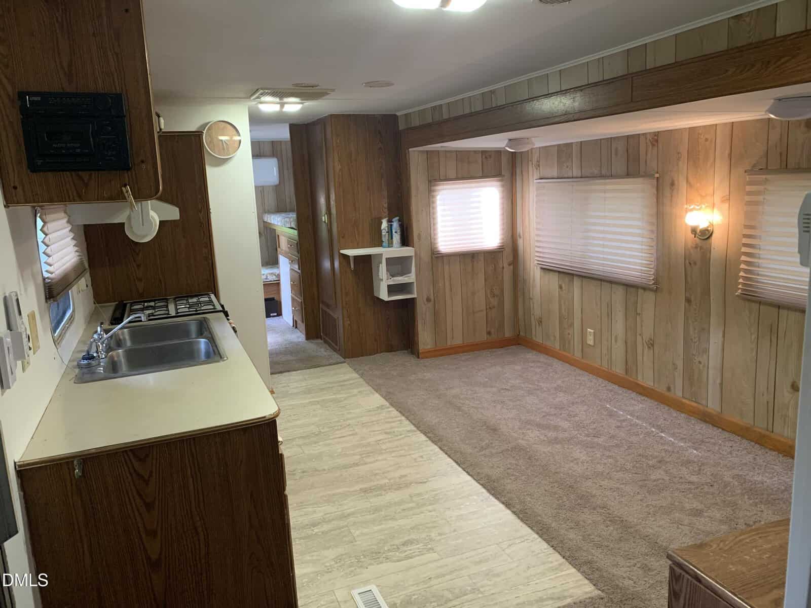 Interior view of a mobile home with wood-paneled walls, beige carpet, and linoleum flooring. The kitchen area has a sink, stove, and cabinets. Living area has windows with blinds. A small hallway leads to the back.
