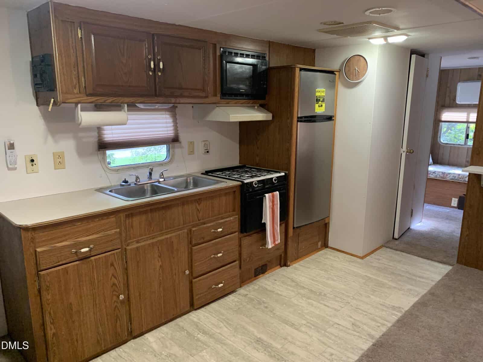 Kitchen area in a mobile home with wood cabinets, a double sink, a gas stove with oven, a microwave, and a small stainless steel refrigerator. A clock is on the wall; a hallway leads to another room.