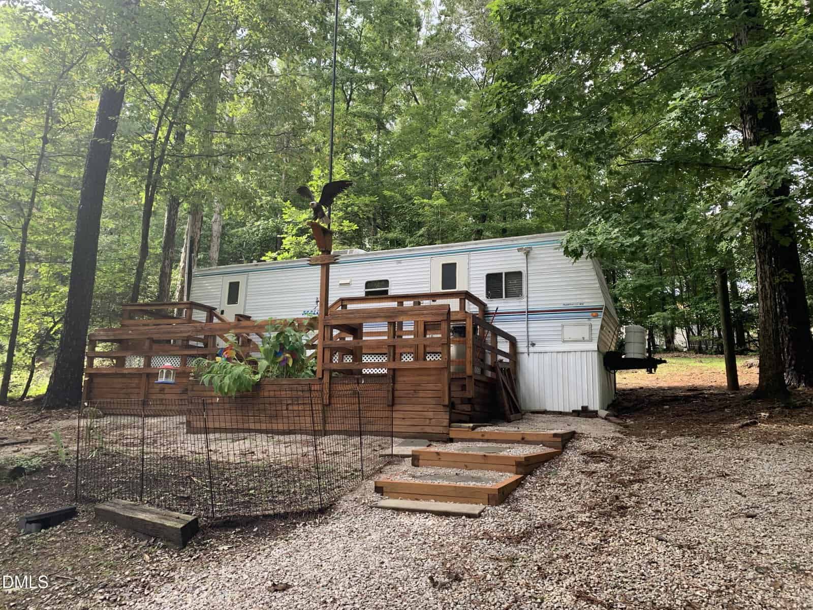 A white trailer sits in a wooded area with a large wooden deck, potted plants, and a small fenced area. Gravel steps lead up to the deck, surrounded by green trees.