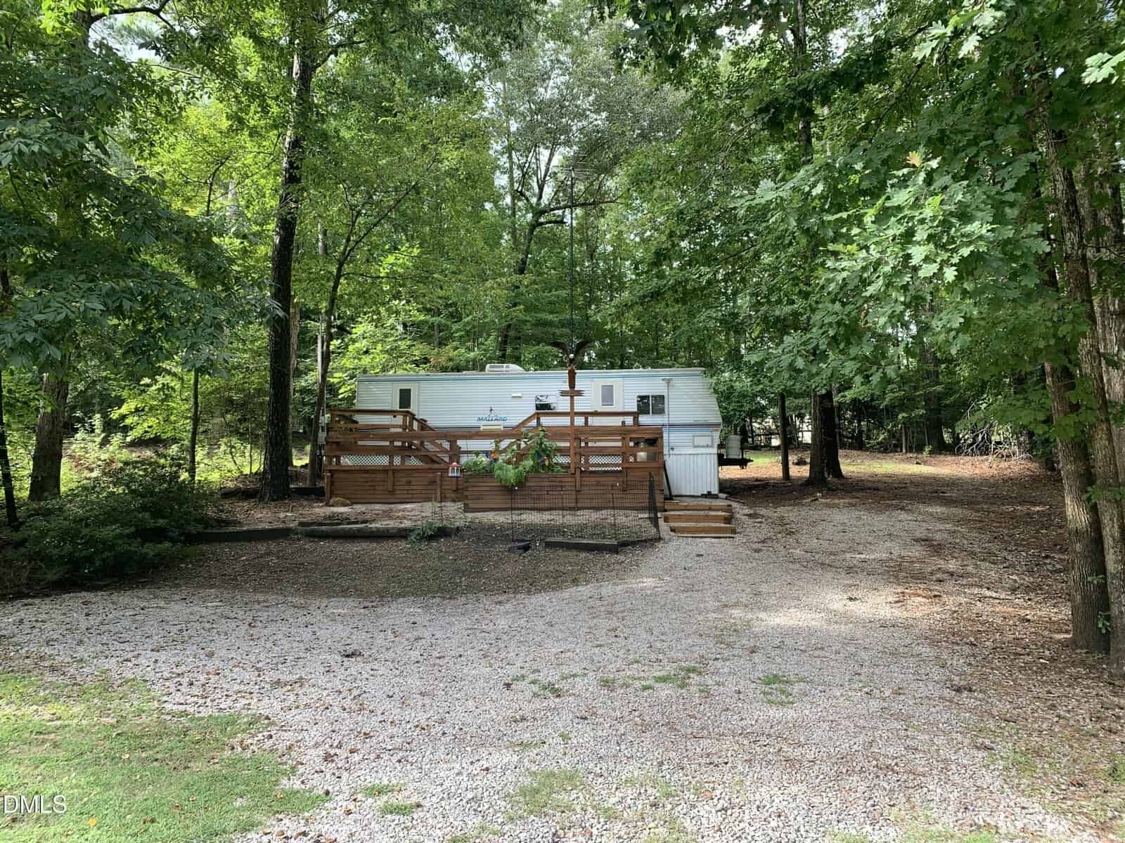 A white travel trailer with a wooden deck is parked among tall trees. The ground is covered with gravel and leaves, and there is a flower planter on the deck railing. The setting is shaded and surrounded by forest.