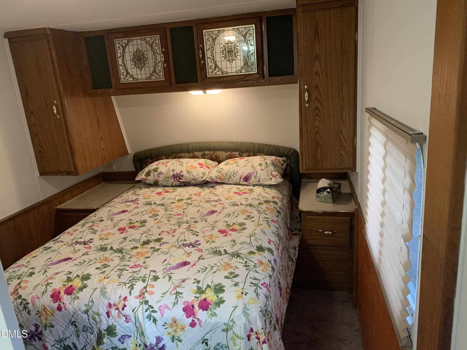 A cozy bedroom with a double bed, floral bedding, wooden cabinets above and beside the bed, small nightstands, and a window with white blinds. Warm lighting gives the space a welcoming feel.