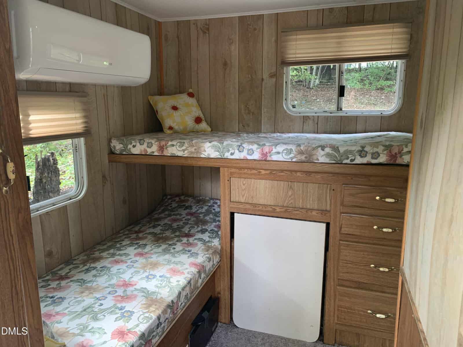 Small bedroom with wooden walls, featuring bunk beds with floral bedding, a pillow with flowers, drawers, a small window with blinds, and an air conditioning unit mounted on the wall.