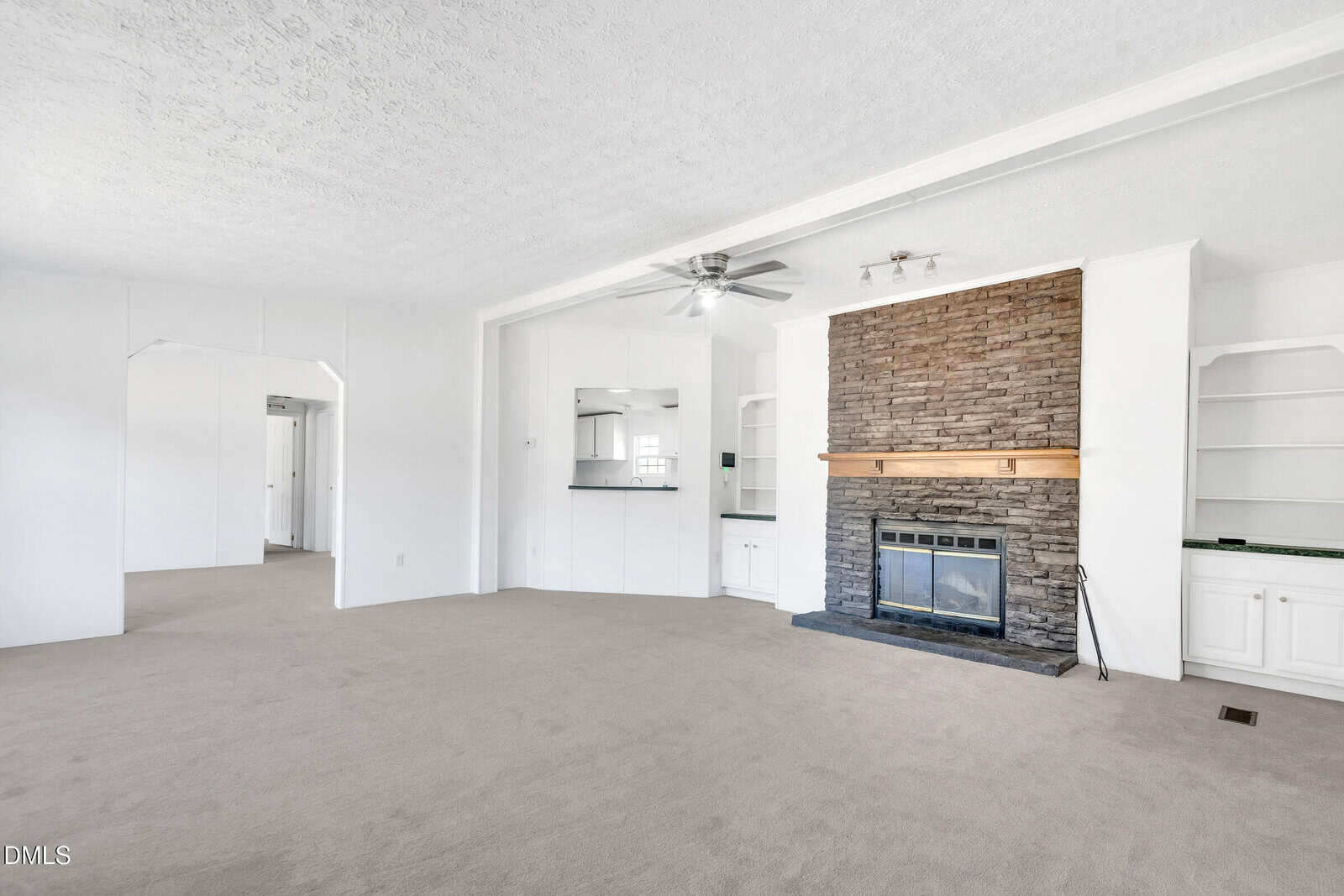 A spacious living room with light-colored carpet, a ceiling fan, a stone fireplace with a wooden mantel, built-in shelves and cabinets, and an open view into an adjacent room and kitchen area.