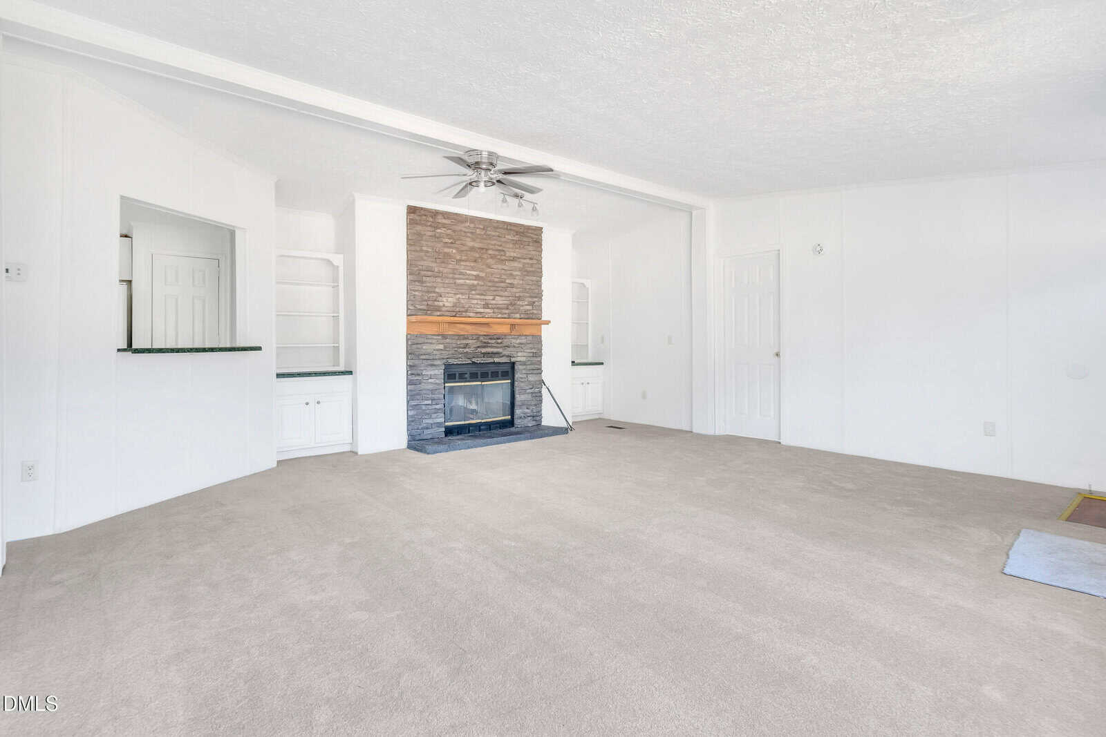 Spacious living room with light beige carpet, white walls, a ceiling fan, built-in shelves, and a stone fireplace with a wooden mantel. Two doorways and windows with green ledges are also visible.
