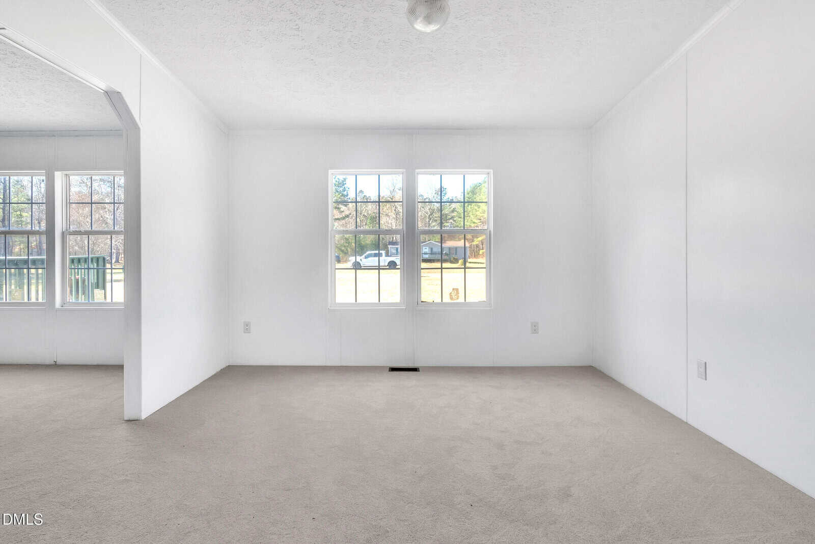 An empty room with white walls, beige carpet, two large windows letting in natural light, and a view of trees and a street outside. An open doorway leads to another area on the left side.