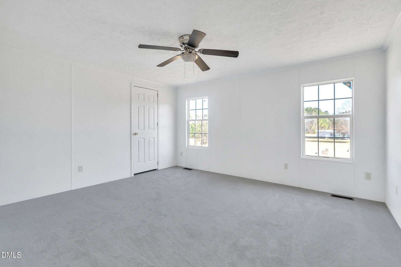 A bright, empty room with white walls, light gray carpet, two windows, a white door, and a ceiling fan with lights. Sunlight enters through the windows, casting light on the carpeted floor.