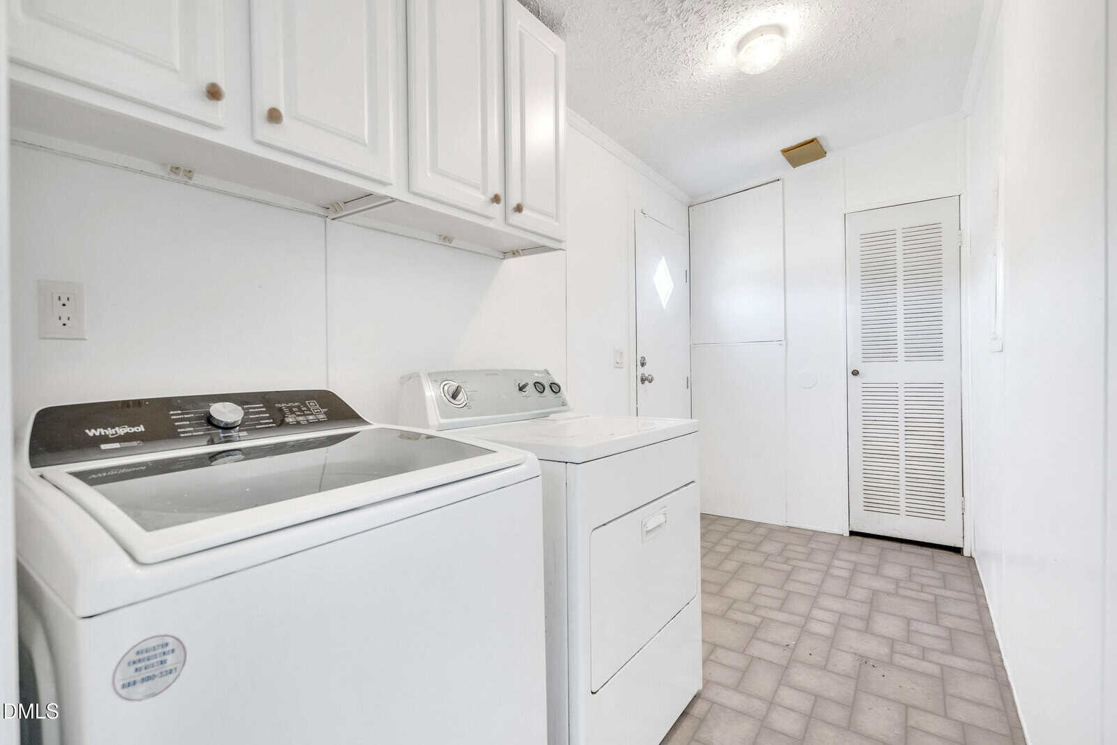A white laundry room with a washing machine, dryer, upper cabinets, tiled floor, and a closed louvered door in the background.