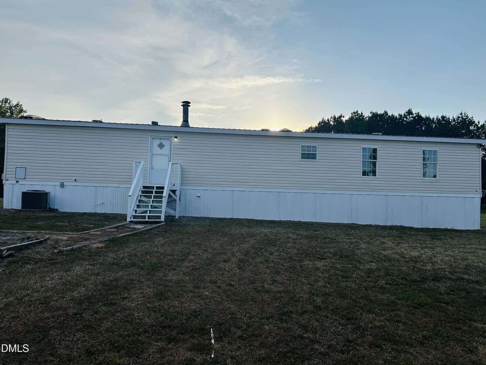 A white single-wide mobile home with a small staircase leading to the front door, several rectangular windows, and a large grassy yard, photographed at sunset.