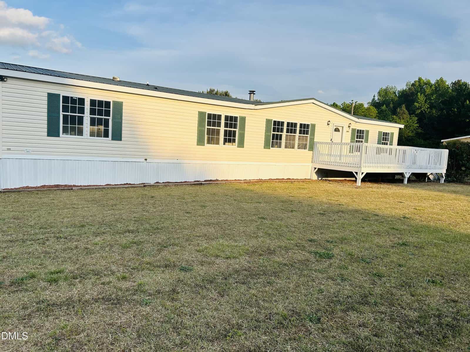 A single-story, light-colored manufactured home with green shutters and a large front deck, set on a grassy yard under a partly cloudy sky. Trees are visible in the background.