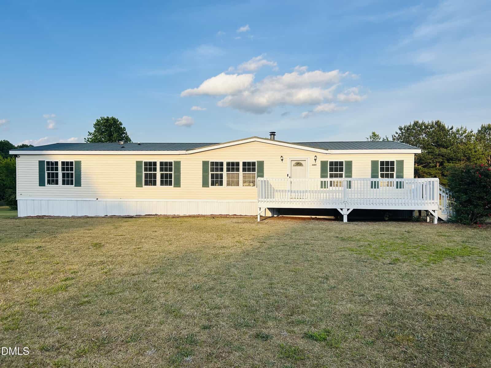 A single-story, light yellow manufactured home with green roof, white trim, and a large white front deck sits on a dry, grassy yard under a blue sky with a few clouds. Trees border the property.